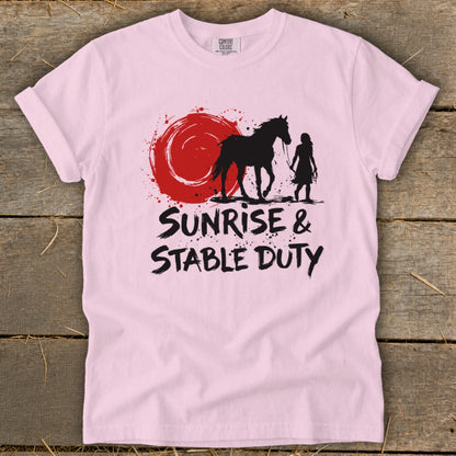 Pink t-shirt with black graphic of horse, rider, and red sun, text "Sunrise & Stable Duty".