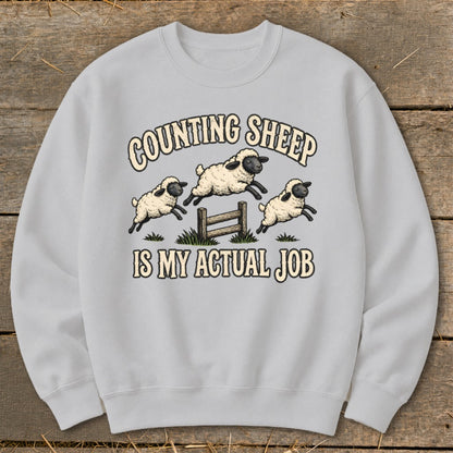 Light gray sweatshirt with three jumping sheep over a fence and text "Counting Sheep is My Actual Job".