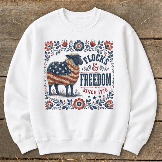 White sweatshirt with a sheep adorned in the American flag, surrounded by floral border. Text reads "Flocks & Freedom Since 1776".