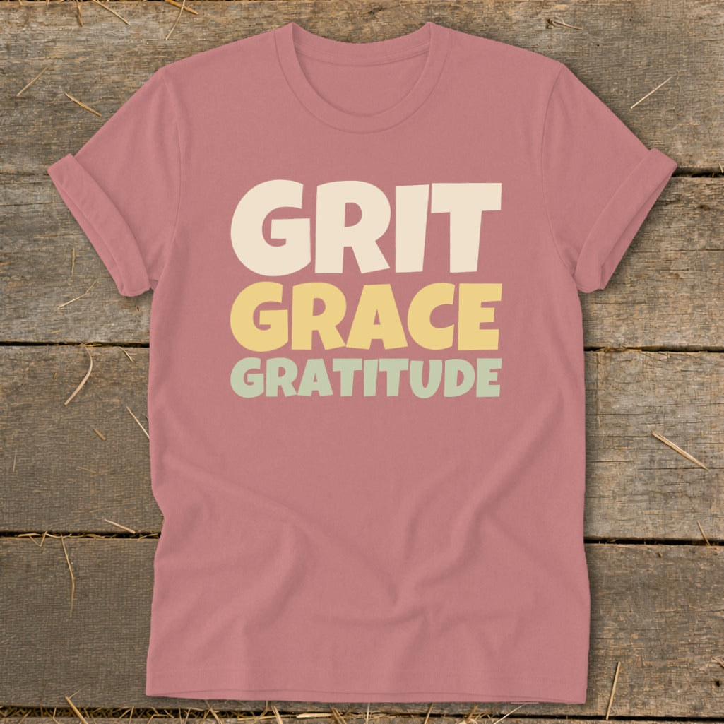 Dusty pink short-sleeve crewneck t-shirt with stacked text reading GRIT GRACE GRATITUDE centered on a wooden background.