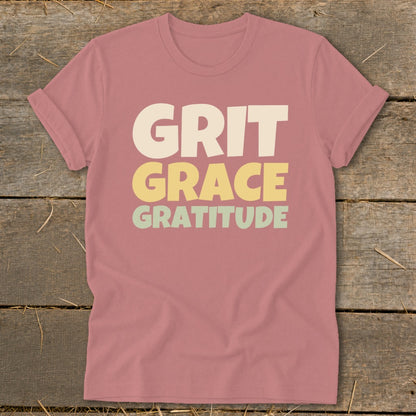 Dusty pink short-sleeve crewneck t-shirt with stacked text reading GRIT GRACE GRATITUDE centered on a wooden background.