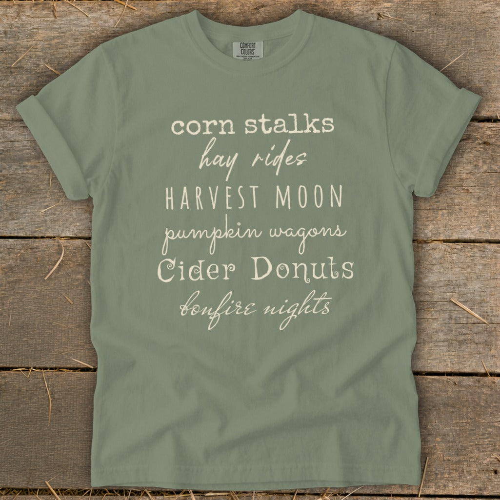 Olive t-shirt with text: corn stalks, hay rides, HARVEST MOON, pumpkin wagons, Cider Donuts, bonfire nights.