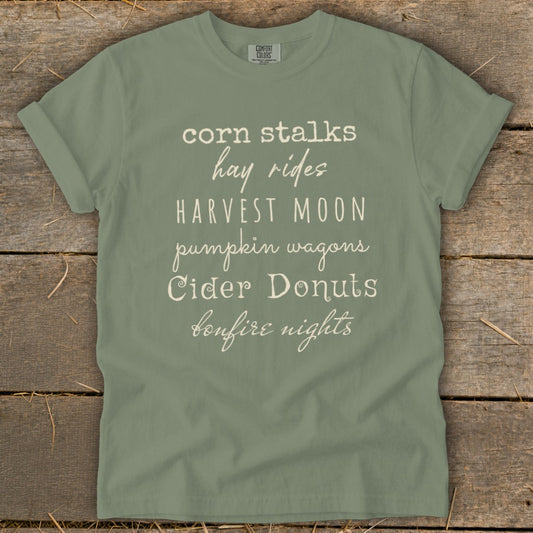 Olive t-shirt with text: corn stalks, hay rides, HARVEST MOON, pumpkin wagons, Cider Donuts, bonfire nights.