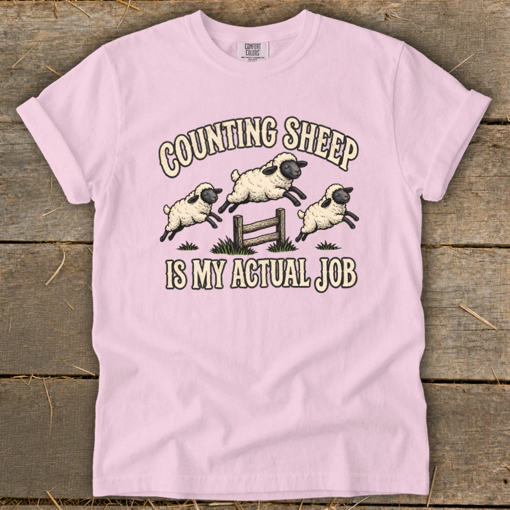 Pink t-shirt with illustration of sheep jumping over a fence with text "Counting Sheep Is My Actual Job".