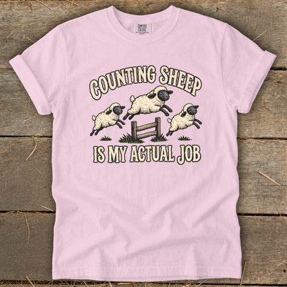 Pink t-shirt with illustration of sheep jumping over a fence with text "Counting Sheep Is My Actual Job".