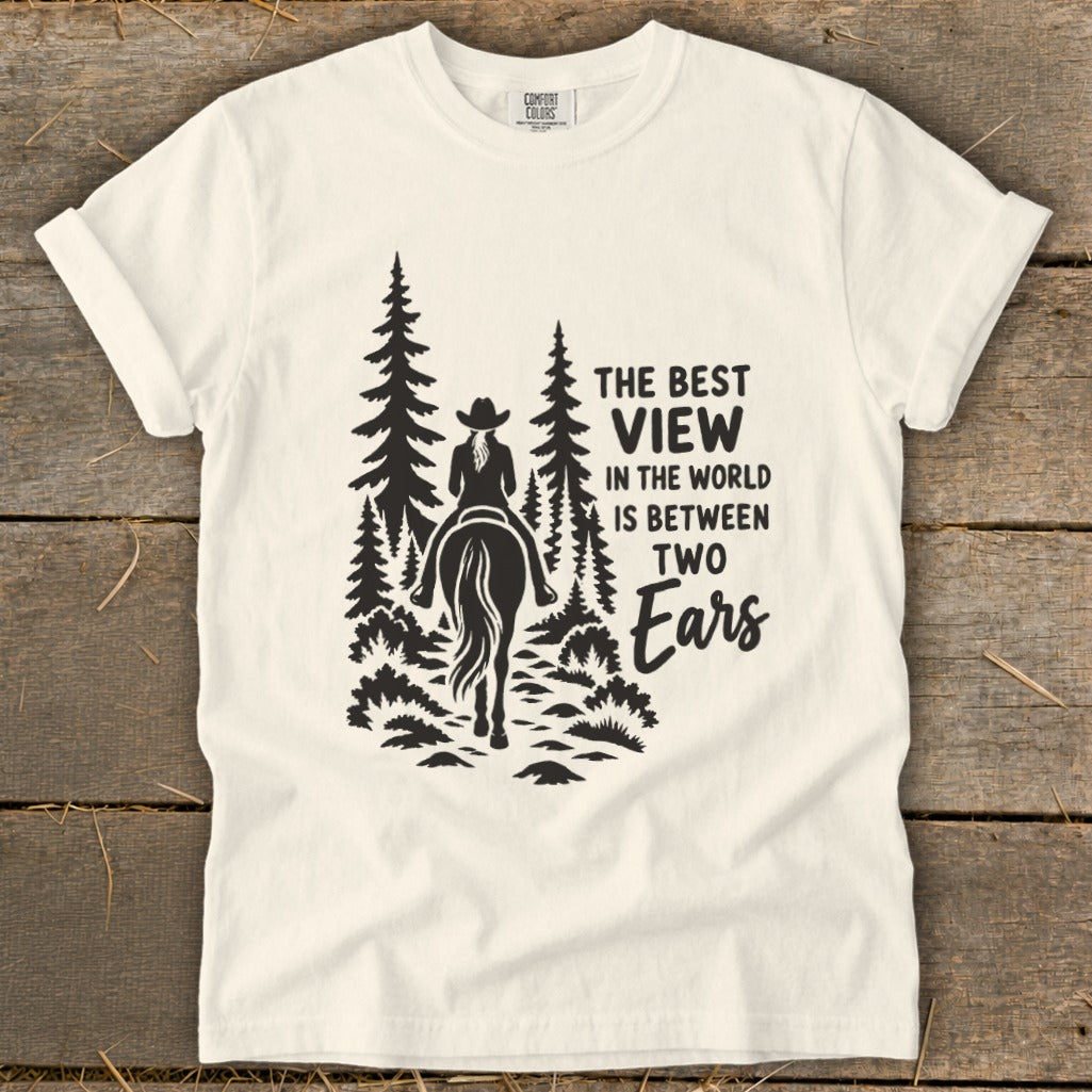 A white crewneck t-shirt featuring a black graphic of a horseback rider in a forest with text reading The Best View in the World is Between Two Ears.