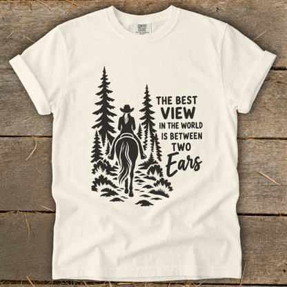A white crewneck t-shirt featuring a black graphic of a horseback rider in a forest with text reading The Best View in the World is Between Two Ears.