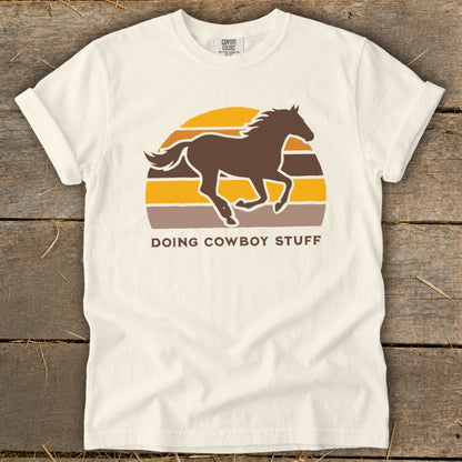 Cream t-shirt with a brown horse running in front of a striped sunset and text "DOING COWBOY STUFF".