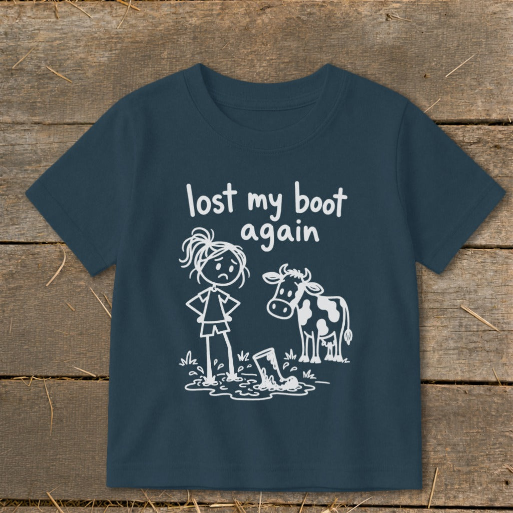 A dark blue children's t-shirt featuring white line art of a sad girl, a cow, and a boot stuck in mud, with the text "lost my boot again."