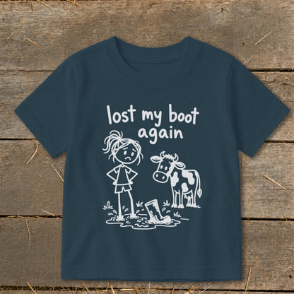A dark blue children's t-shirt featuring white line art of a sad girl, a cow, and a boot stuck in mud, with the text "lost my boot again."