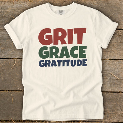 Off-white t-shirt with rolled sleeves laid flat, featuring stacked text reading "GRIT, GRACE, GRATITUDE" in red, green, and blue.