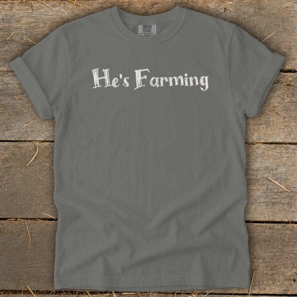 Gray t-shirt with white text "He's Farming" on a rustic wooden background.
