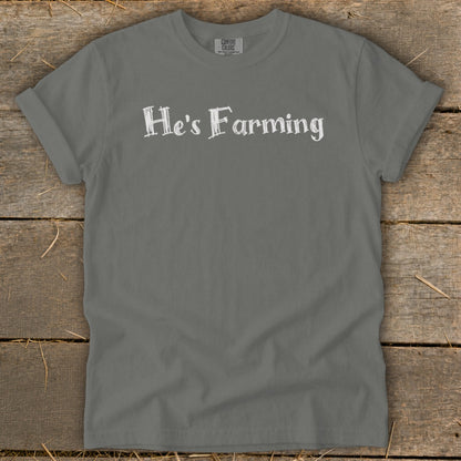 Gray t-shirt with white text "He's Farming" on a rustic wooden background.