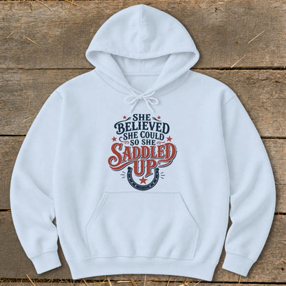 White hoodie with "She believed she could so she saddled up" text and horseshoe graphic.
