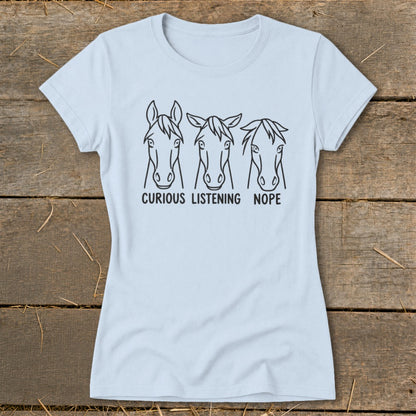 A light blue crewneck t-shirt featuring three horse head illustrations with the text "Curious, Listening, Nope" below them, laid flat on a rustic wooden background.