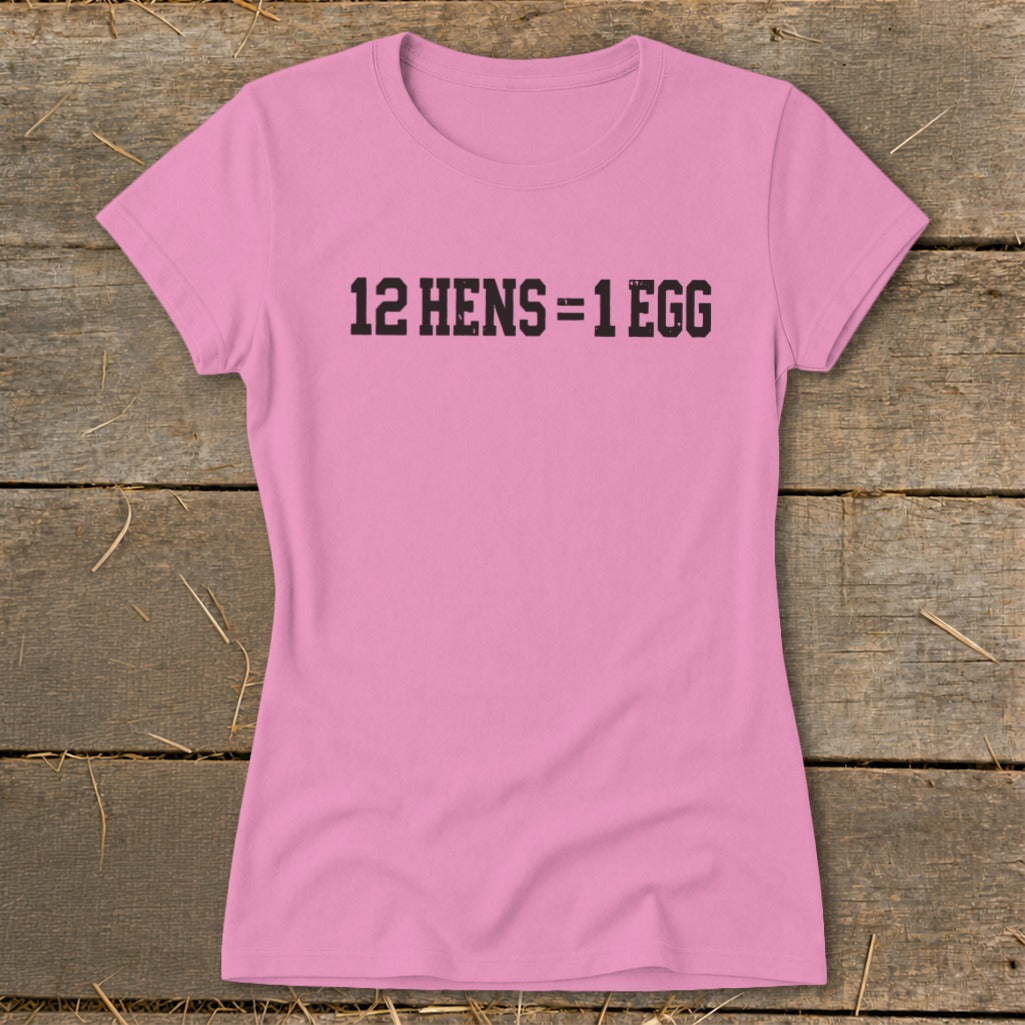 A pink women's t-shirt rests on a rustic wooden background, featuring black text that reads 12 HENS = 1 EGG.