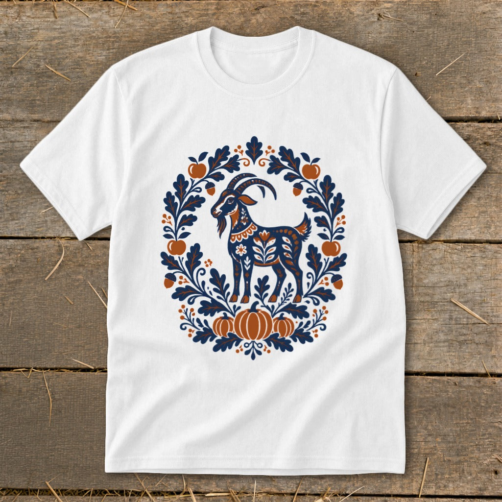 A white short-sleeved t-shirt features a folk-art graphic of a goat surrounded by an autumn wreath of leaves, pumpkins, and apples.