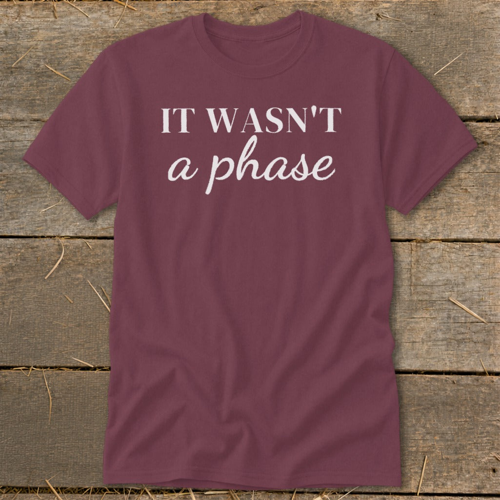 Maroon t-shirt with white text "IT WASN'T a phase" on a wooden background.