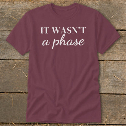 Maroon t-shirt with white text "IT WASN'T a phase" on a wooden background.