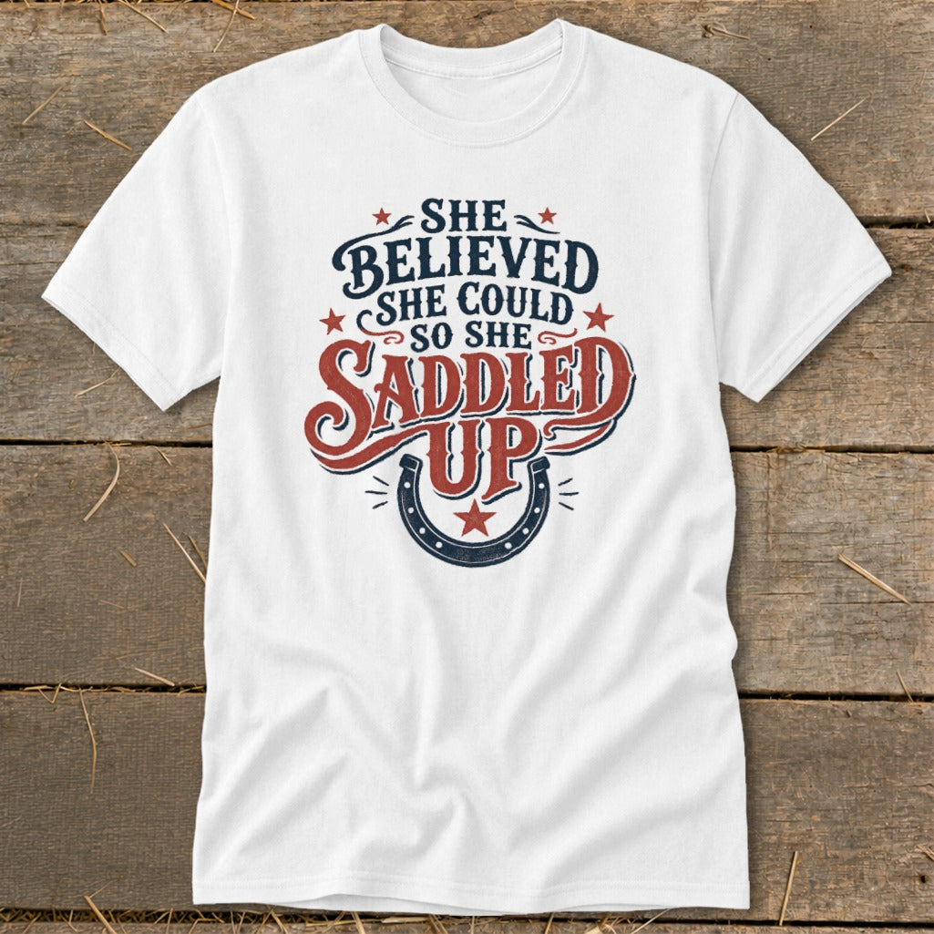 White t-shirt with "She believed she could so she saddled up" text and horseshoe graphic.