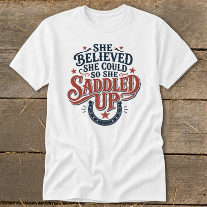 White t-shirt with "She believed she could so she saddled up" text and horseshoe graphic.