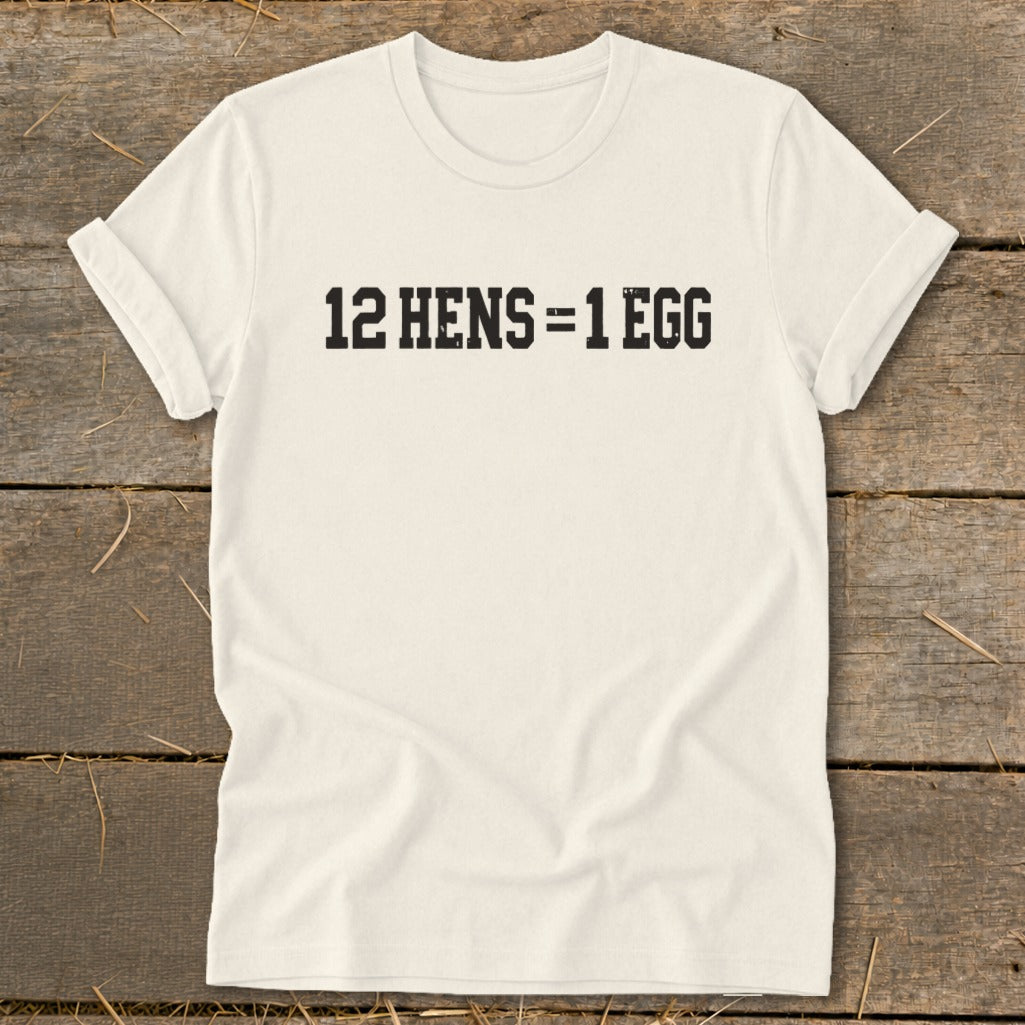 A light beige crewneck t-shirt with black text that reads 12 HENS = 1 EGG, laid flat on a rustic wooden background.