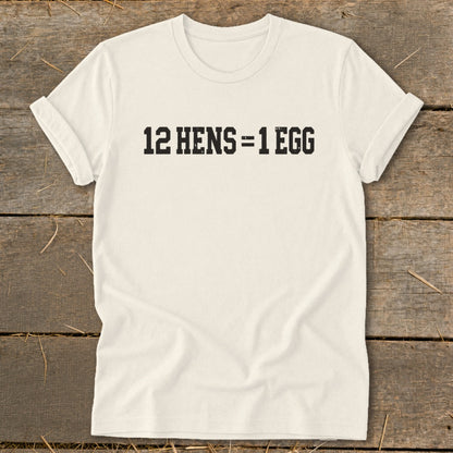 A light beige crewneck t-shirt with black text that reads 12 HENS = 1 EGG, laid flat on a rustic wooden background.