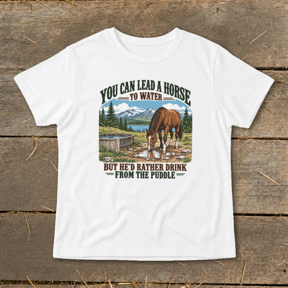 A white t-shirt featuring a graphic of a horse drinking from a puddle with mountains and the text You Can Lead A Horse To Water But He'd Rather Drink From The Puddle.