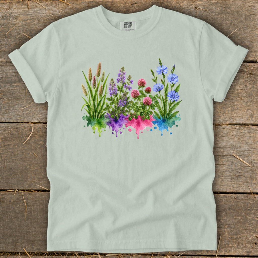 A light sage green t-shirt laid flat on a rustic wooden background, featuring a colorful watercolor wildflower graphic.