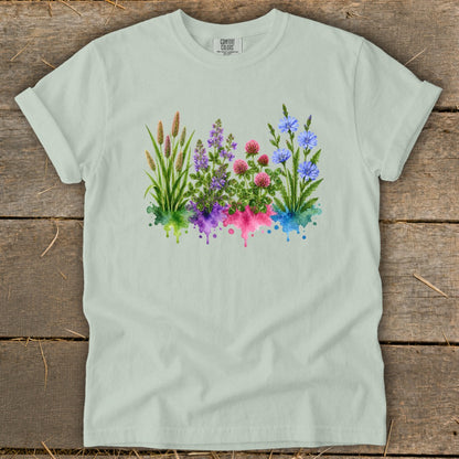 A light sage green t-shirt laid flat on a rustic wooden background, featuring a colorful watercolor wildflower graphic.