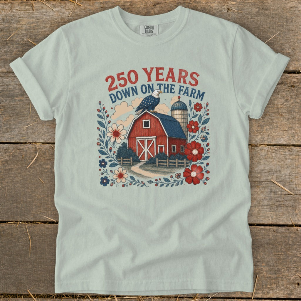 Light blue t-shirt graphic with red barn, eagle, silo, and patriotic flowers. Text reads: "250 Years Down On The Farm".