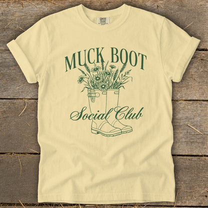 Yellow t-shirt with green text and graphic of muck boots filled with flowers.