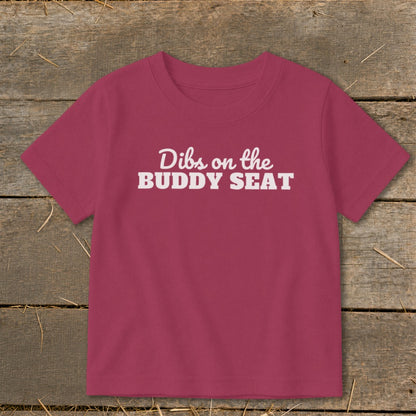 Toddler t-shirt with white text "Dibs on the BUDDY SEAT" on maroon fabric.