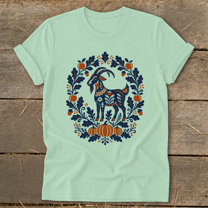 Light green t-shirt with a folk-art design of a blue goat inside an autumn wreath.