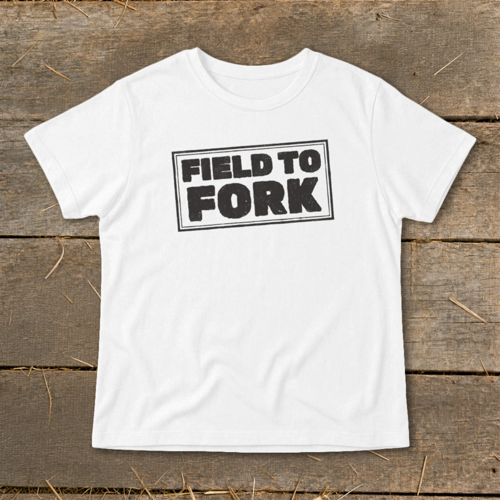 A white short-sleeved t-shirt with a distressed black graphic reading FIELD TO FORK centered on a wooden background.