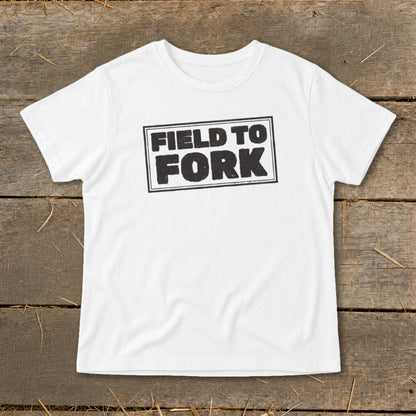 A white short-sleeved t-shirt with a distressed black graphic reading FIELD TO FORK centered on a wooden background.