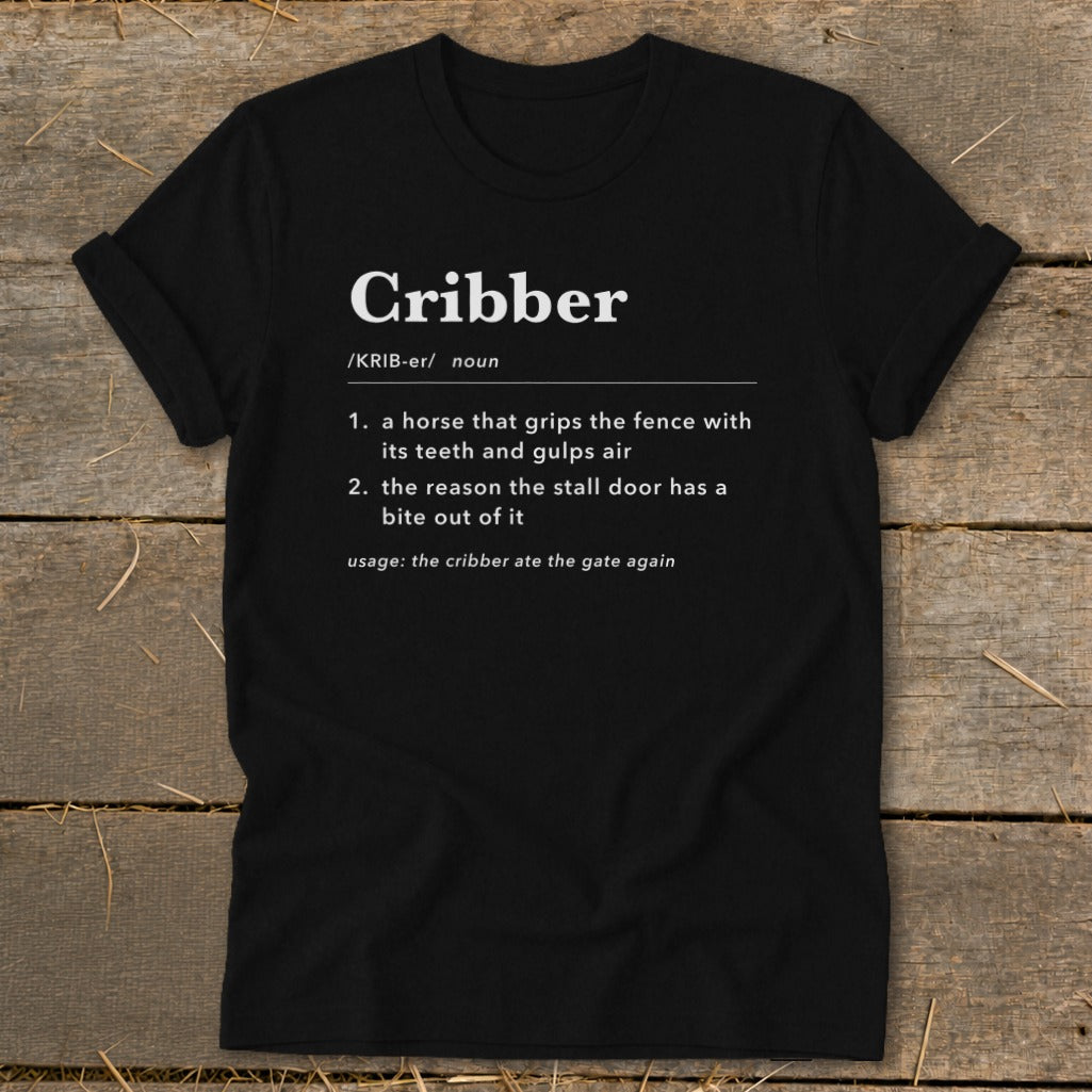 Black t-shirt featuring dictionary-style text defining the word Cribber and laying flat on wood.