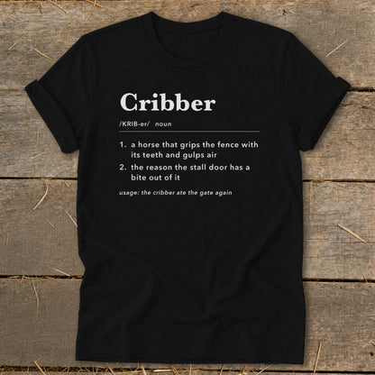 Black t-shirt featuring dictionary-style text defining the word Cribber and laying flat on wood.