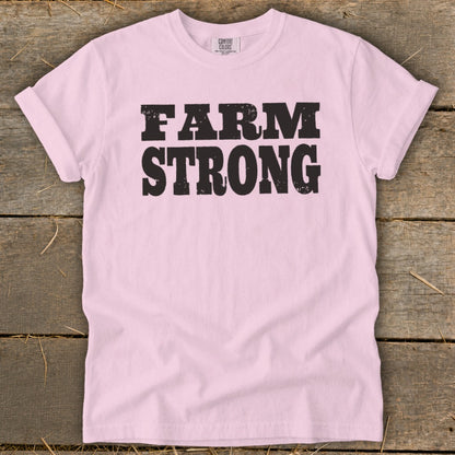 Light pink t-shirt with rolled sleeves featuring the bold black text FARM STRONG on a wooden background.