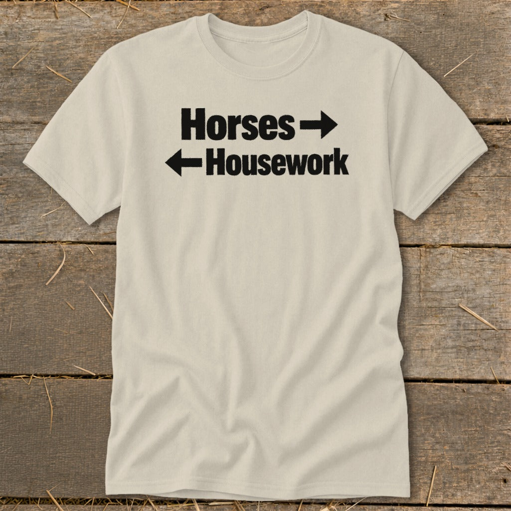 A cream-colored crewneck t-shirt with black text that reads Horses with a right arrow above Housework with a left arrow.