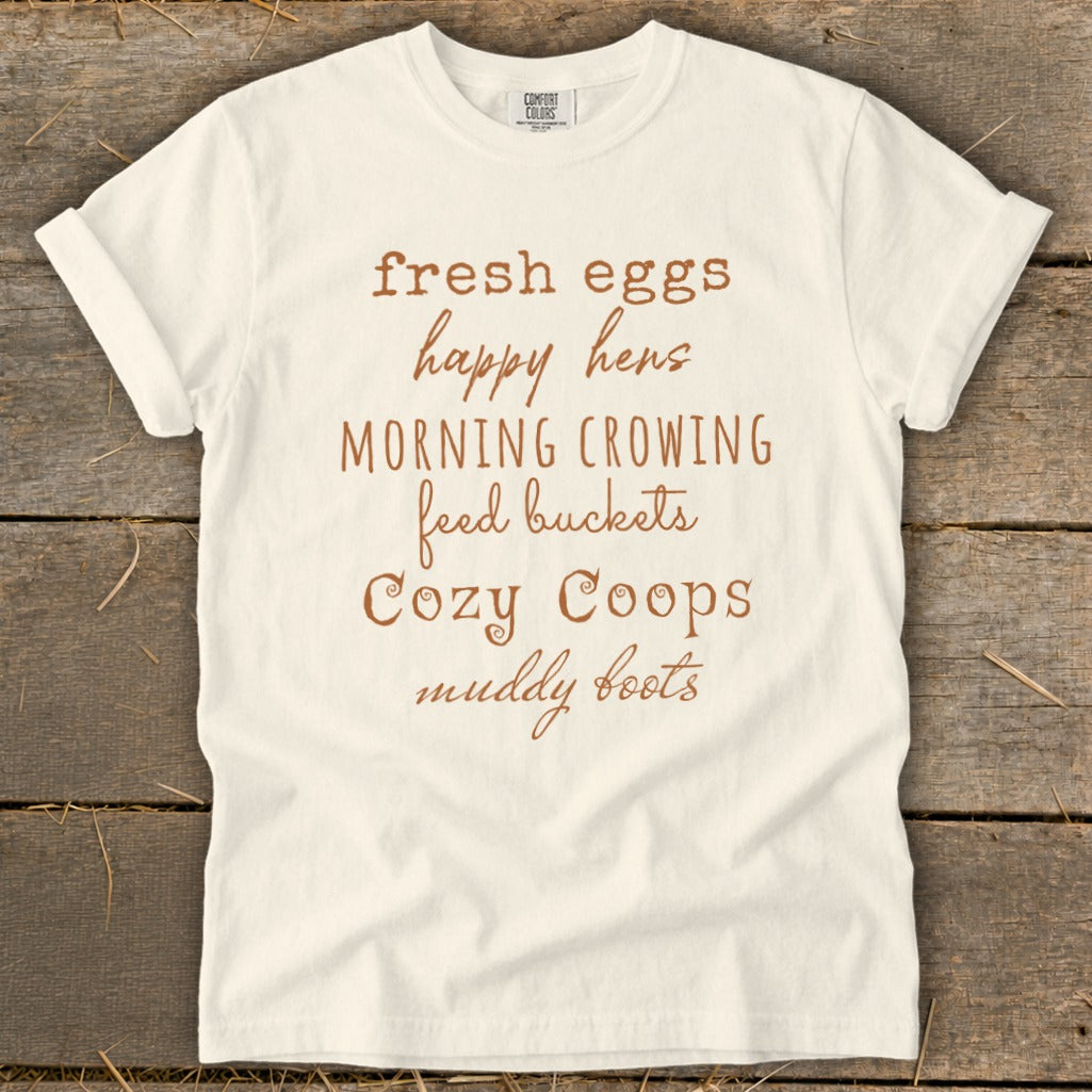 White t-shirt with brown text reading "fresh eggs, happy hens, morning crowing, feed buckets, Cozy Coops, muddy boots".