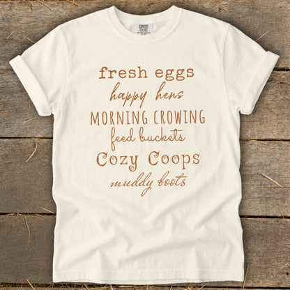 White t-shirt with brown text reading "fresh eggs, happy hens, morning crowing, feed buckets, Cozy Coops, muddy boots".