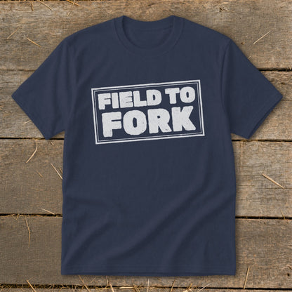 A dark blue t-shirt with a white graphic reading "Field to Fork" inside a rectangular border, laid flat on a rustic wooden surface.