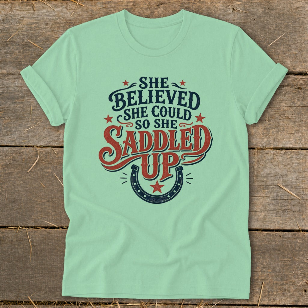 Light green t-shirt with "She believed she could so she saddled up" text and horseshoe.