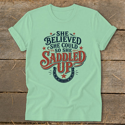 Light green t-shirt with "She believed she could so she saddled up" text and horseshoe.
