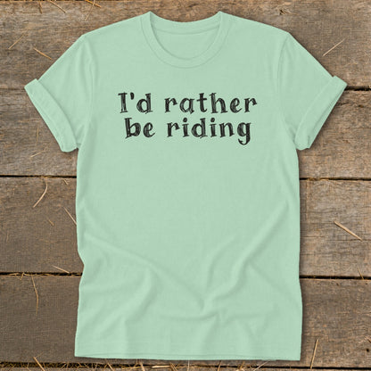 Light green t-shirt with "I'd rather be riding" text on a wooden background.