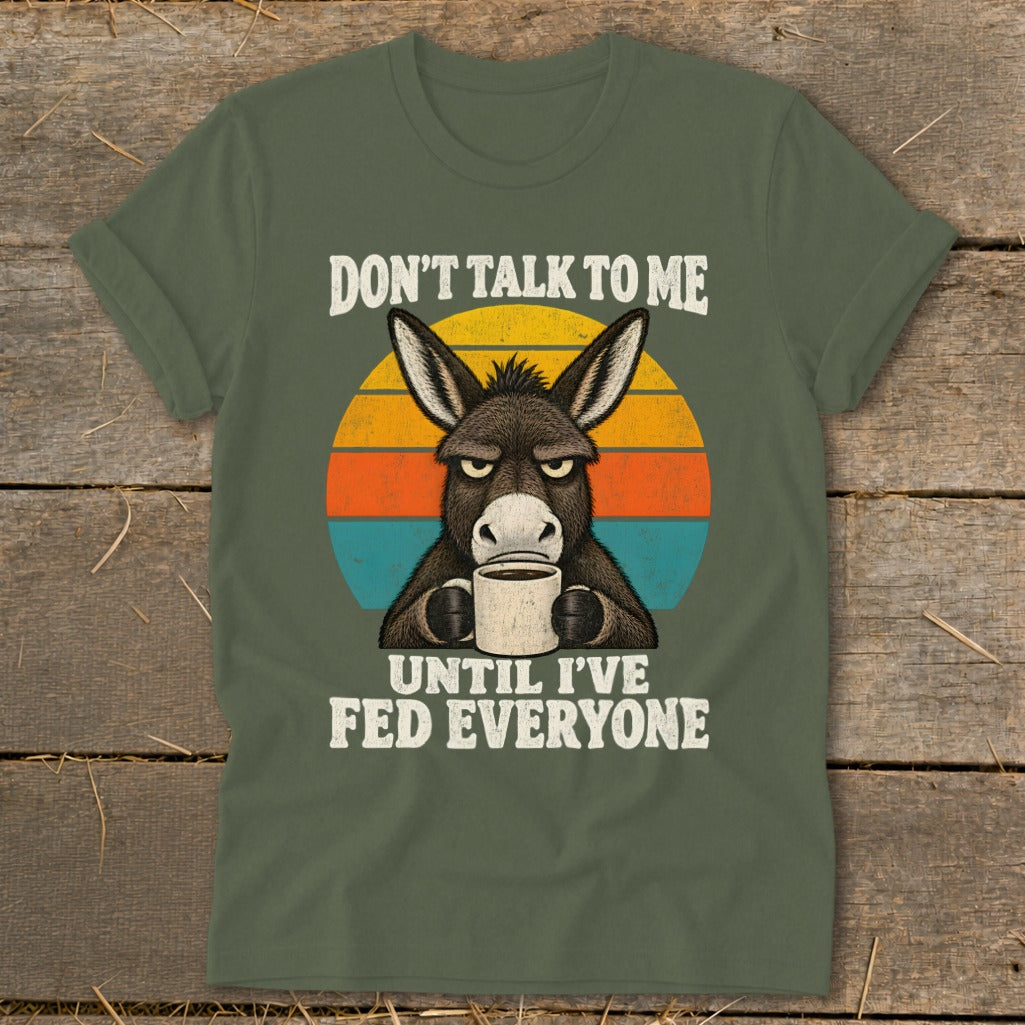 An olive green t-shirt featuring a grumpy donkey holding a coffee mug with the text Don't Talk to Me Until I've Fed Everyone against a sunset background.