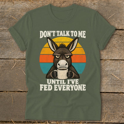 An olive green t-shirt featuring a grumpy donkey holding a coffee mug with the text Don't Talk to Me Until I've Fed Everyone against a sunset background.