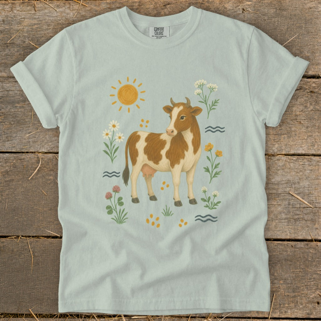 Brown and white cow surrounded by flowers, sun, and waves on light blue t-shirt.