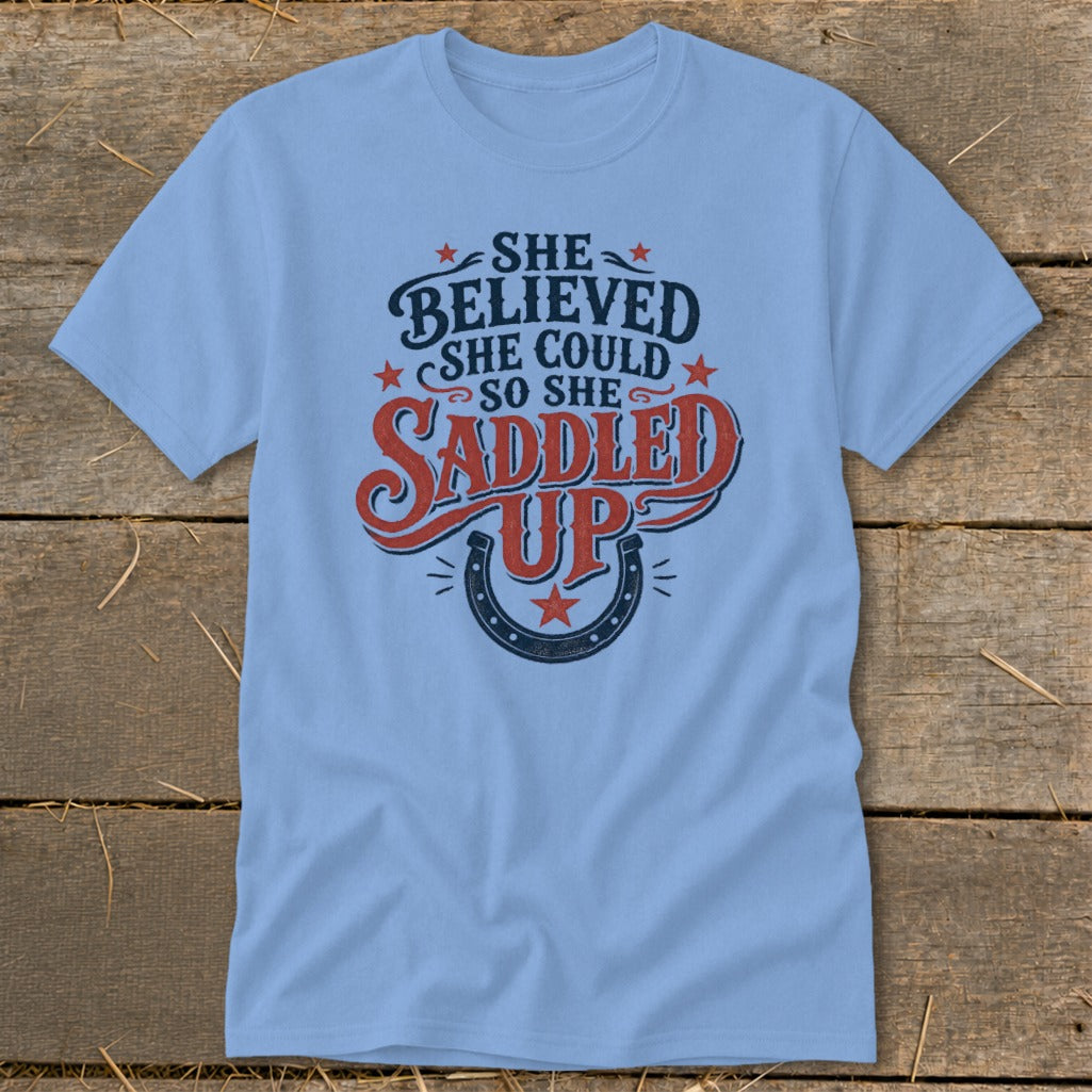 Light blue t-shirt with "She believed she could so she saddled up" text and horseshoe.